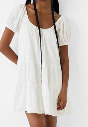 Vrouw met lange vlechten, gekleed in een korte, witte jurk met laagjes, pofmouwen en een ronde halslijn.