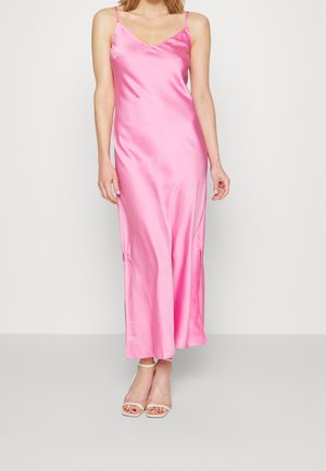 Mujer que lleva un vestido tipo slip de satén rosa, sin mangas y largo hasta el tobillo, con tirantes finos, y tacones beige abiertos en los dedos, posando contra un fondo liso.