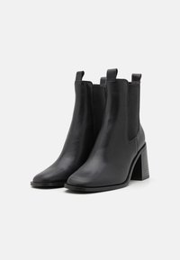 Paire de bottines en cuir noir avec talons carrés, panneaux latéraux élastiques et languettes de tirage, présentées sur un fond blanc.