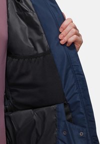 Das Innere einer marineblauen und schwarzen Jacke zeigt eine Netz- und glänzende Fütterung. Eine Hand hält die Jacke geöffnet und offenbart ihren Stoff und die Struktur.