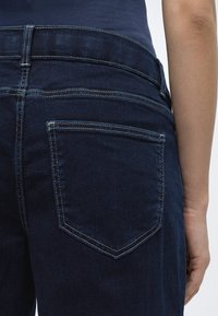 Πλάγια όψη πίσω μέρους ατόμου που φοράει σκοτεινό μπλε τζιν και ναυτικό μπλουζάκι, εστιάζοντας στην πίσω δεξιά τσέπη και το χέρι, σε απλό φόντο.