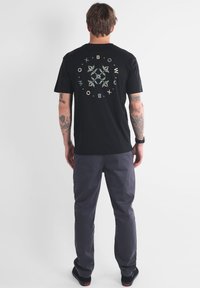 T-shirt en coton noir avec un graphisme circulaire représentant des feuilles et du texte en couleurs claires au dos, associé à un pantalon gris et des chaussures noires.