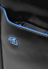 Bolso de cuero negro con una textura suave, con acentos azules a lo largo de los bordes y un logo metálico azul. Cierre de cremallera visible.