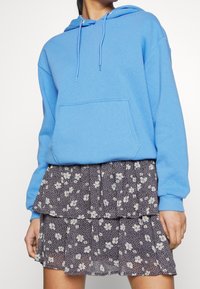 Personne portant un sweat à capuche bleu clair avec poche avant, associé à une mini-jupe noire à fleurs superposées.