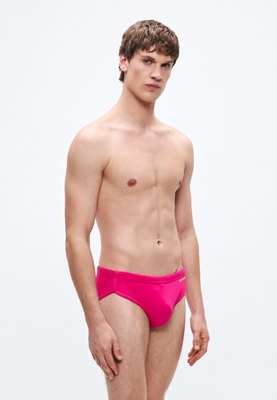 Ροζ ανδρικό μαγιό σε γυαλιστερό υλικό, με χαμηλή μέση και ελάχιστη κάλυψη. Ομαλή υφή με διακριτική λεπτομέρεια λογότυπου.