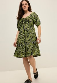 Grünes und schwarzes gemustertes Kleid mit kurzen Puffärmeln, einem quadratischen Ausschnitt und einem Bindeband vorne. Getragen mit flachen schwarzen Schuhen.