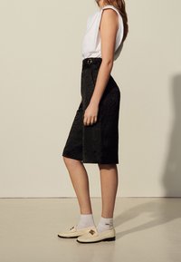 Jupe noire longueur genou avec motif ondulé texturé, associée à un haut blanc sans manches et des mocassins beige, agrémentée d'un accent décoratif en métal.