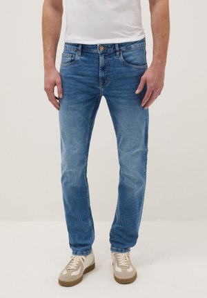 Man die lichtblauwe slim-fit spijkerbroek, witte sneakers en een wit T-shirt draagt, staand tegen een effen achtergrond.