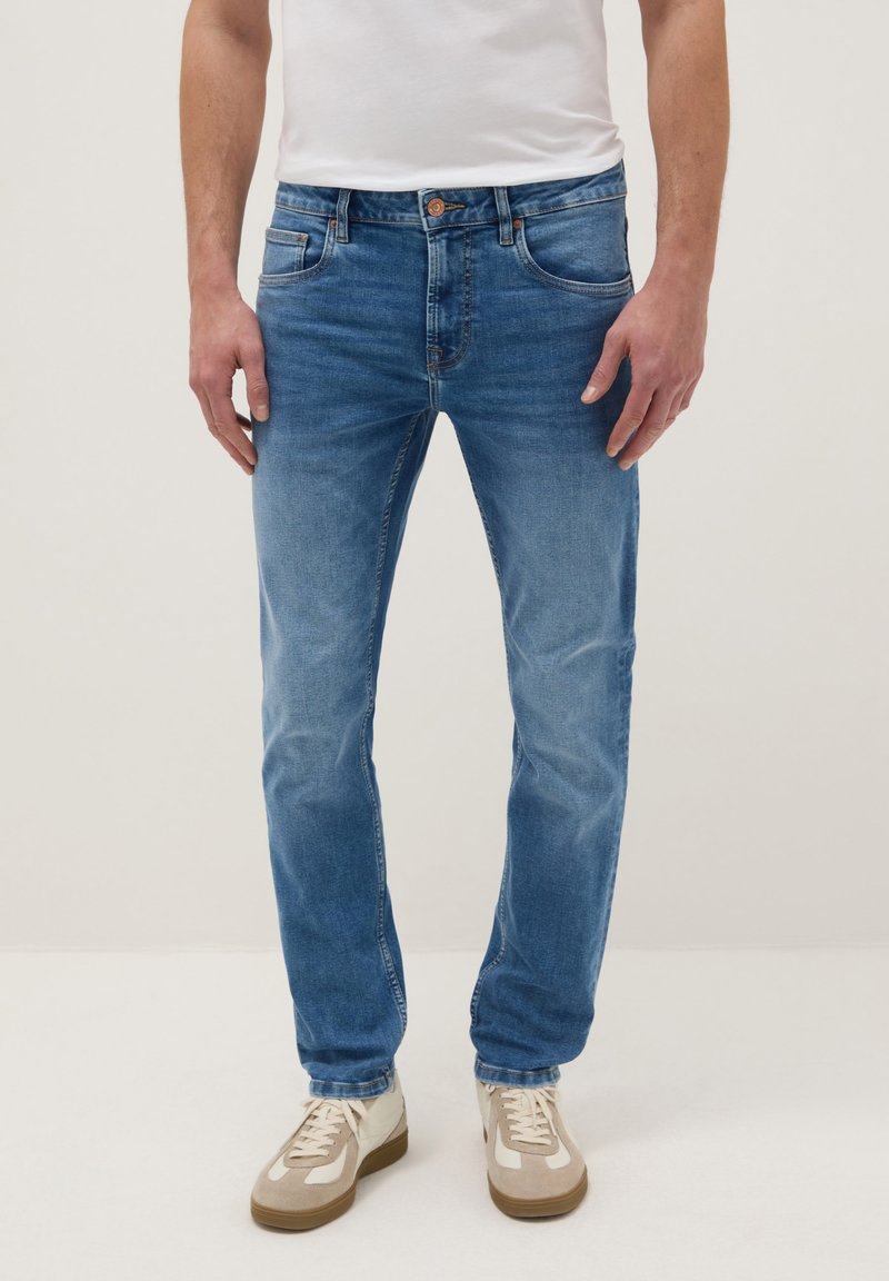 Mann trägt hellblaue Slim-Fit-Jeans, weiße Sneakers und ein weißes T-Shirt, steht vor einem einfarbigen Hintergrund.