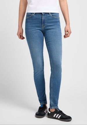 Femme portant un jean slim bleu moyen, des baskets noires Adidas et une chemise blanche, debout contre un fond clair uni.