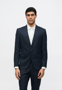 Tengerészkék öltöny két gombbal és felvágott hajtókával, fehér inggel párosítva. Testhez álló szabás és sima anyag, oldalzsebekkel és mintás textillel.