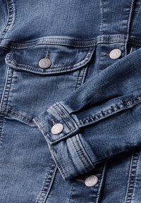 Giacca di jeans di blu medio. Caratterizzata da bottoni argentati, tasche sul petto e doppie cuciture. Tessuto liscio con leggerissime aree sbiadite.