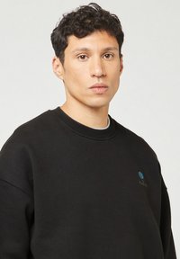 Felpa nera realizzata in misto cotone, con colletto a girocollo, spalle scese e un piccolo logo blu sul petto. Texture liscia.