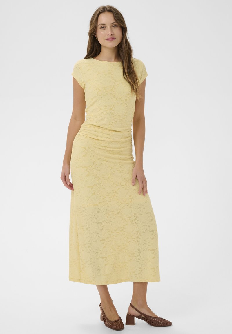 Femme vêtue d'un haut en dentelle jaune à manches courtes et d'une jupe longue assortie, portée avec des chaussures à talons blocs tressés marron, debout devant un fond blanc.