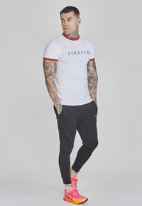 Witte t-shirt met rode rand en zwart "SIKSILK" logo, gecombineerd met zwarte slim-fit joggers en felroze sportschoenen.