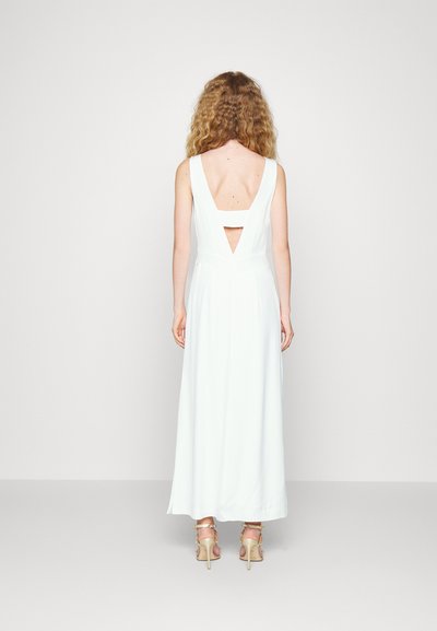 Vestido maxi branco sem mangas com costas em V profundo e detalhes recortados. Tecido suave, forma fluida e design minimalista. Usado com saltos bege.