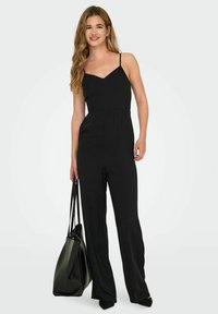 Zwarte jumpsuit met spaghettibandjes, v-hals, elastische taille en een wijd uitgesneden ontwerp. Gecombineerd met een grote zwarte tote bag.