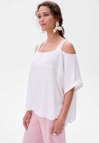 Biała bluzka bez ramiączek z kwadratowym dekoltem i dekoracyjnymi paskami. Charakteryzuje się luźnym krojem i gładką fakturą, zestawiona z różowymi spodniami.