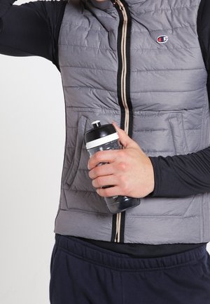 Mężczyzna w szarej pikowanej kamizelce i czarnej koszuli z długim rękawem trzyma czarno-białą butelkę na wodę na jednolitym tle.