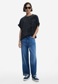 Übergröße schwarzes T-Shirt mit Grafikdruck, blaue weit geschnittene Jeans und blaue Sneakers mit weißen Details. Lässiger Schnitt mit umgeschlagenen Ärmeln.