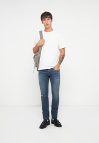 Camiseta blanca, jeans azules desgastados con rasgaduras, mocasines negros y un bolso de hombro gris. Fondo simple, pose de pie.