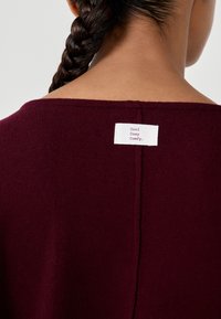 Burgundowy sweter z szerokim dekoltem i prostym szwem na plecach. Metka przy szyi z napisem "Cool, Cosy, Comfy" na białym tle.