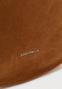 Ruda zomšinė rankinė, pasižyminti lygiu paviršiumi. Prekinis ženklas „Coccinelle“ subtiliai įspaustas sidabro spalva. Apvalaus formos su siuvinėtomis kraštinėmis.