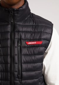 Schwarze Steppweste mit hohem Kragen, horizontalem Steppmuster, Frontreißverschluss und einem roten rechteckigen Aufnäher mit der Aufschrift "JACKIT."