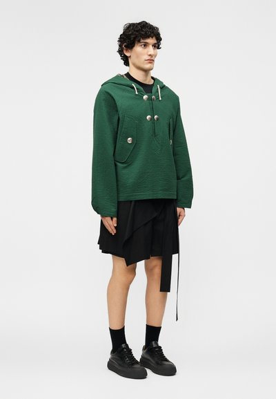 Άνδρας με σγουρά μαύρα μαλλιά, που φοράει πράσινο μπουφάν με κουκούλα και κουμπιά, μαύρη φούστα με στρώσεις, μαύρες κάλτσες και μαύρα αθλητικά παπούτσια, στέκεται σε λευκό φόντο.