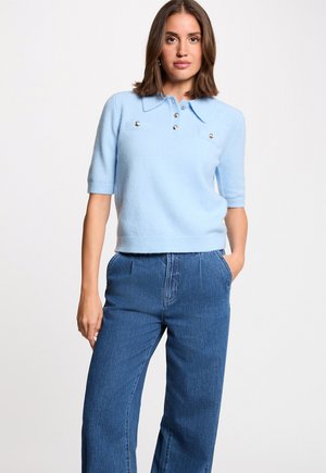 Frau trägt ein hellblaues Strickoberteil mit kurzen Ärmeln, Kragen und Knöpfen, kombiniert mit einer hoch taillierten blauen Jeans, steht mit einer Hand in der Tasche.