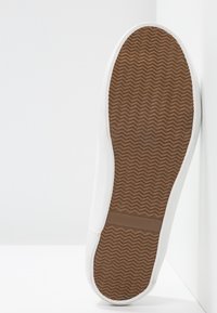Semelle d'une basket blanche montrant une semelle extérieure en caoutchouc marron avec un motif de bande en zigzag et une bande de grip horizontale traversant la voûte plantaire.