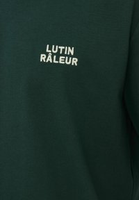 Tissu vert foncé avec texte brodé en blanc "LUTIN RÂLEUR" centré sur la partie supérieure du vêtement.