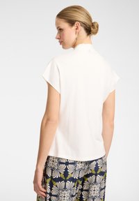 Top bianco a maniche corte, con collo alto e texture liscia. Indossato con pantaloni blu navy a fantasia, con motivi floreali e di ananas.