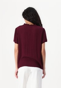 Femme aux longs cheveux foncés portant un chemisier bordeaux à manches courtes avec une texture subtile et un pantalon blanc, vue de dos sur fond blanc.