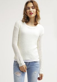Pull en tricot blanc avec un col rond, des manches longues et des poignets côtelés. Associé à un jean déchiré bleu clair.