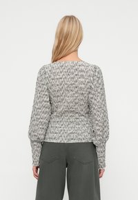 Ξανθή γυναίκα που φοράει μια υφασμάτινη, μακριά μανίκια γκρίζα και λευκή μπλούζα με σκούρες γκρι παντελόνες, απεικονιζόμενη από πίσω σε απλό φόντο.