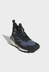 Musta ja laivansiniset Adidas Terrex vaelluskengät, joissa on valkoinen pehmustettu pohja, mustat nauhat ja vetovaljaat kielellä ja kantapäällä harmaata taustaa vasten.