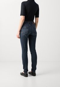 Skinny jeans in dark blue denim, featuring a mid-rise waist, five-pocket design, and a brown leather patch on the back waistband.