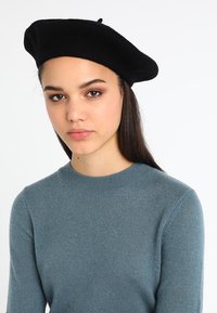 Um boina de lã preta repousa na cabeça. O modelo usa um suéter de malha de manga longa azul claro com um decote redondo e uma textura suave.