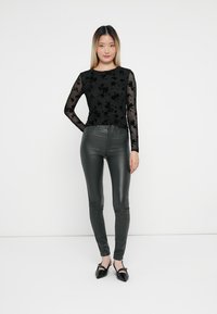 Sort langærmet top med blomstrede blondedetaljer parret med tætsiddende mørkegrønne leggings og sorte spidse sko. Glat tekstur overalt.