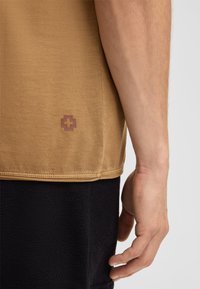 Korte mouwen bruine T-shirt van zachte stof, met een klein rood Zwitsers kruislogo vlakbij de zoom en een gestructureerd zwart kledingstuk eronder.