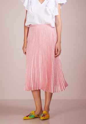 Kvinde iført en hvid bluse med flæser, en pink plissee midi-nederdel og gule slip-on sko med farverigt blomsterbroderi, stående på et beige gulv.