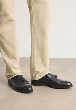 Mocassini in pelle nera con pompon, profilo basso, punta rettangolare, abbinati a pantaloni beige chiaro a gamba dritta. Texture liscia, design minimale.