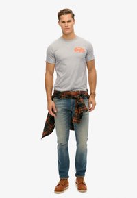 Grijze katoen T-shirt met oranje tekst, blauwe jeans, bruine leren schoenen en een geruite flanellen shirt geknoopt om de taille.