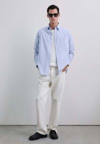 Camicia a righe blu chiaro con bottoni sopra un maglione bianco, abbinata a pantaloni bianchi larghi e scarpe slip-on nere. Occhiali da sole neri inclusi.
