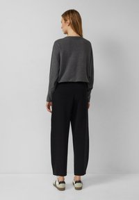 Une femme portant un pull à côtes gris foncé, un pantalon noir ample et des baskets blanches se tient face à un mur gris clair.