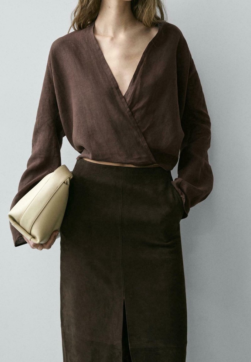 Femme portant un chemisier cache-cœur brun foncé et une jupe assortie, tenant une pochette beige sur un fond clair uni.