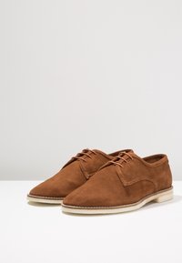 Chaussures en cuir suédé marron à bout pointu, dotées d’une fermeture à lacets et d’une semelle de couleur claire. Texture lisse avec des accents de couture courbée.