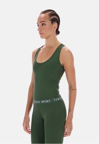 Zöld sportos ujjatlan felső, lekerekített nyakkivágással és testhezálló szabással. Texturált anyag jellemzi, és kontrasztos logós pánt található a derékon.