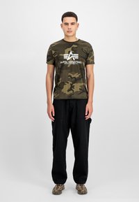 T-shirt καμουφλάζ σε σκούρο πράσινο και μαύρο με λευκό λογότυπο, συνδυασμένο με μαύρο cargo παντελόνι και παπούτσια με σχέδιο. Χαλαρή εφαρμογή, κοντά μανίκια.